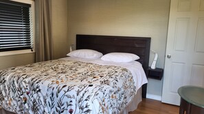 A neatly made bed with a floral bedspread, a bedside table with a lamp, and a door with a handle. - River Heights Motel (Courtenay)