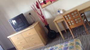 A room with a bed, a wooden dresser, a television on top, a small table with a chair, and a vase with red sticks. - Hotel Coronado (Ensenada)