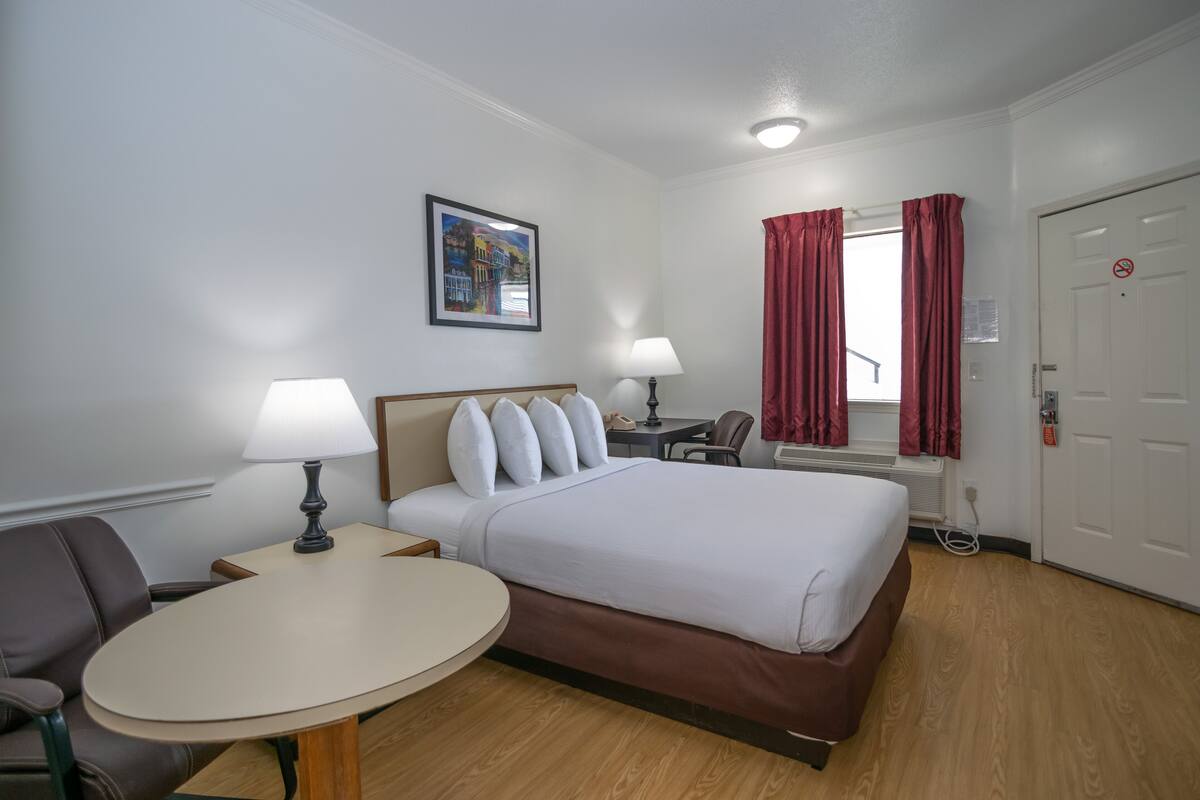 A hotel room with a bed, a round table, two lamps, a desk, a chair, a window with red curtains, and a door with a no-smoking sign.