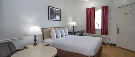 Quarto de hotel com cama, mesa redonda, duas luminárias, escrivaninha, cadeira, janela com cortinas vermelhas e porta com placa de proibido fumar.