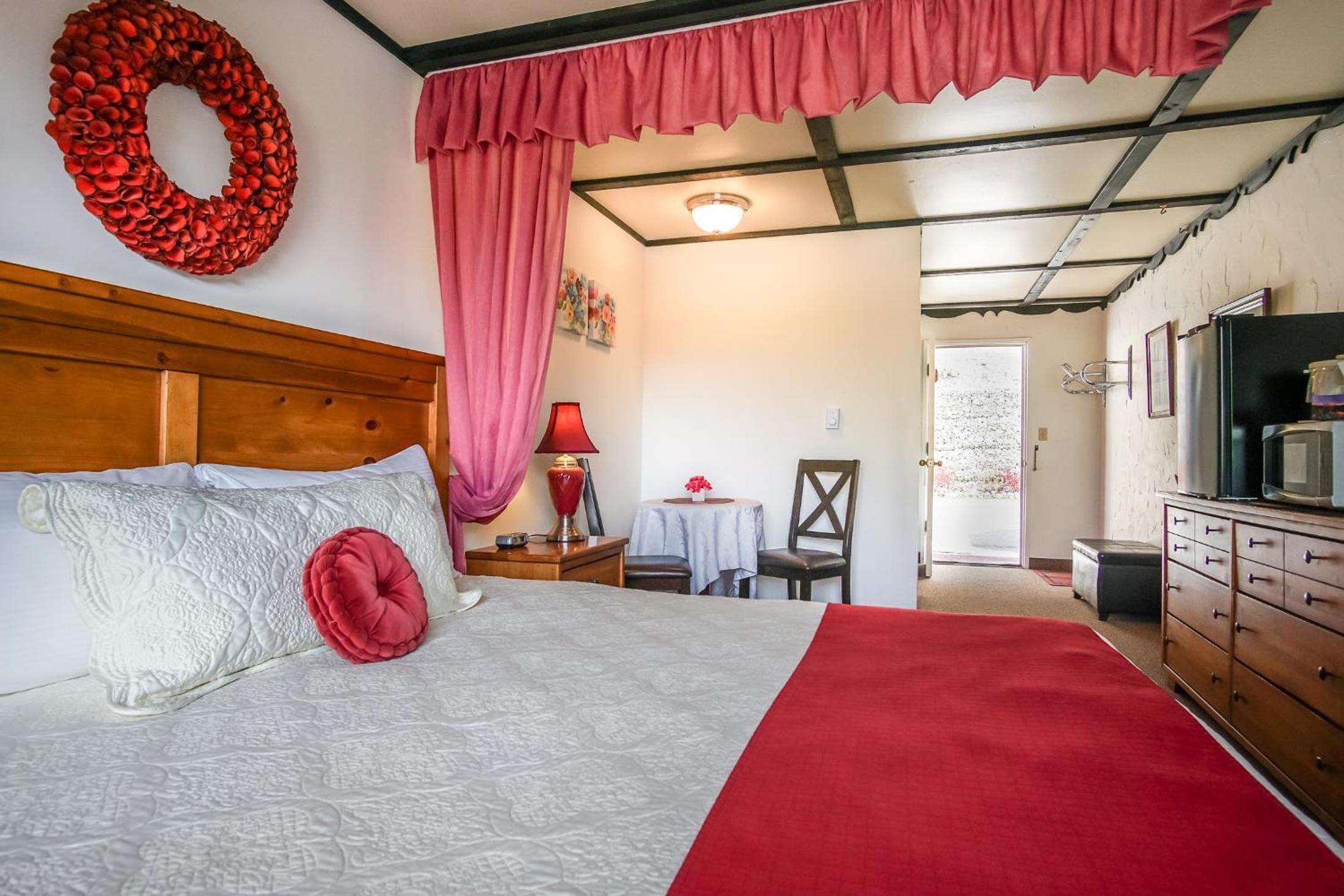 Une chambre avec une tête de lit en bois, un grand lit avec une literie à motifs, un coussin rouge, un rideau rouge, une décoration murale rouge, une petite table avec une lampe et un téléviseur posé sur un meuble en bois.