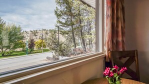 A room with a window overlooking a street and trees, a wooden chair, and a table with a vase of flowers. - Gables Motel (Radium Hot Springs)