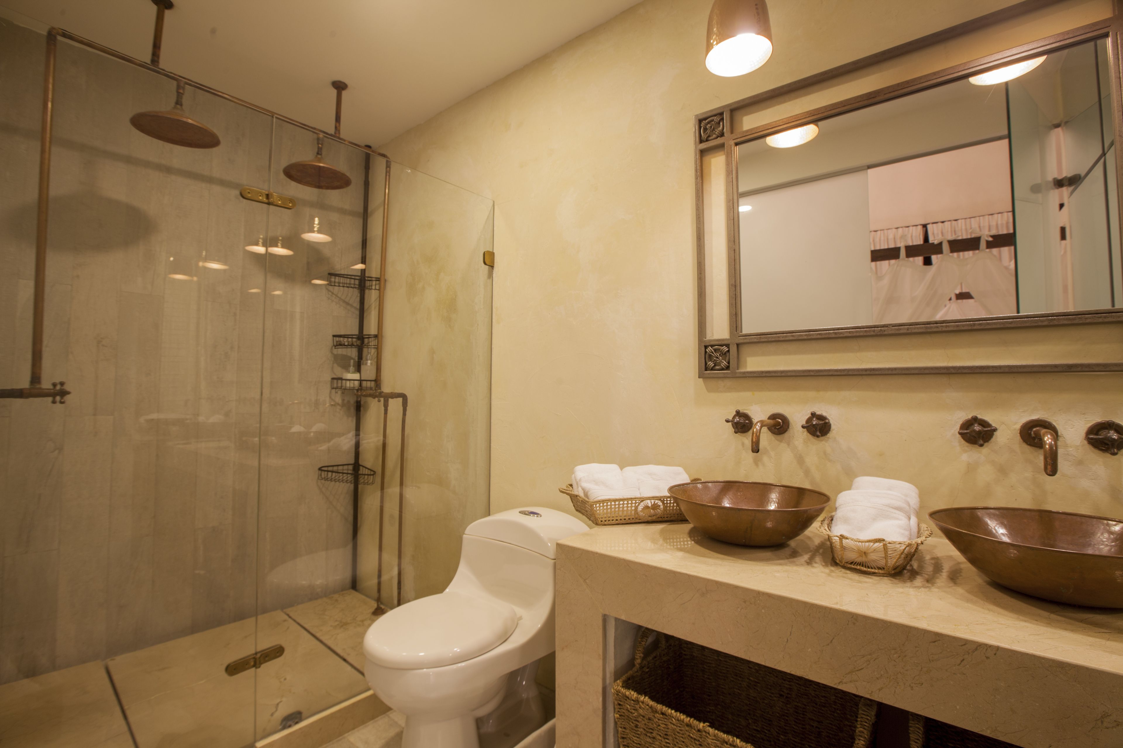 Casa de banho moderna com chuveiro em vidro, sanita branca e lavatório com duas grandes pias de cobre e um espelho acima.