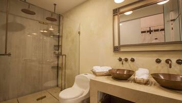 Casa de banho moderna com chuveiro em vidro, sanita branca e lavatório com duas grandes pias de cobre e um espelho acima.