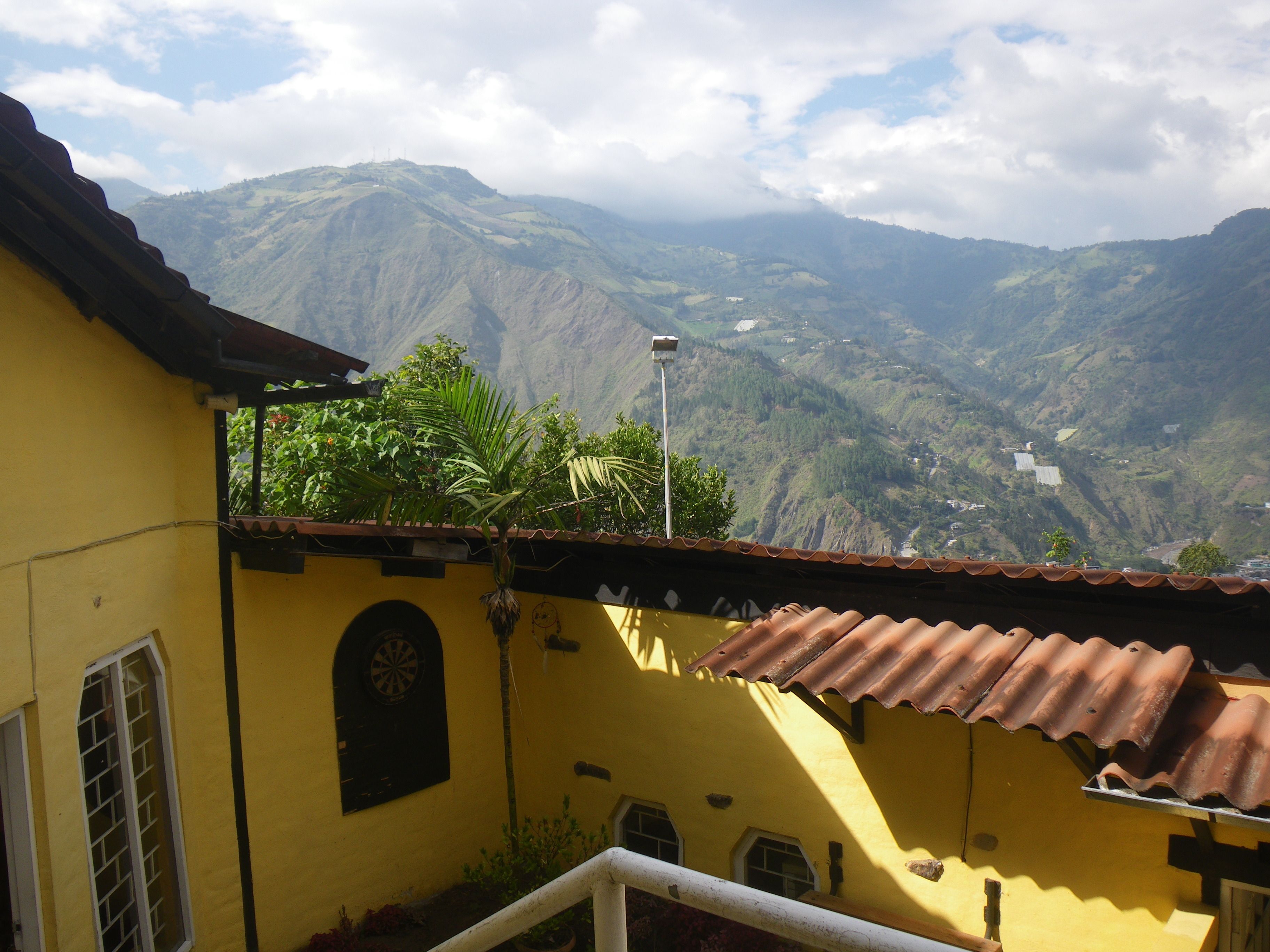 A yellow building with a tiled roof, a small garden, and a view of a valley with mountains in the background.