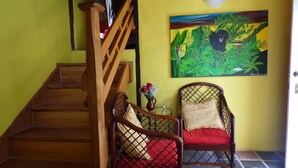 Un escalier mène à une pièce où se trouvent deux fauteuils en osier, l’un avec un coussin rouge et l’autre avec un coussin beige, un tableau représentant un lion au mur et une petite table sur laquelle est posé un vase de fleurs.