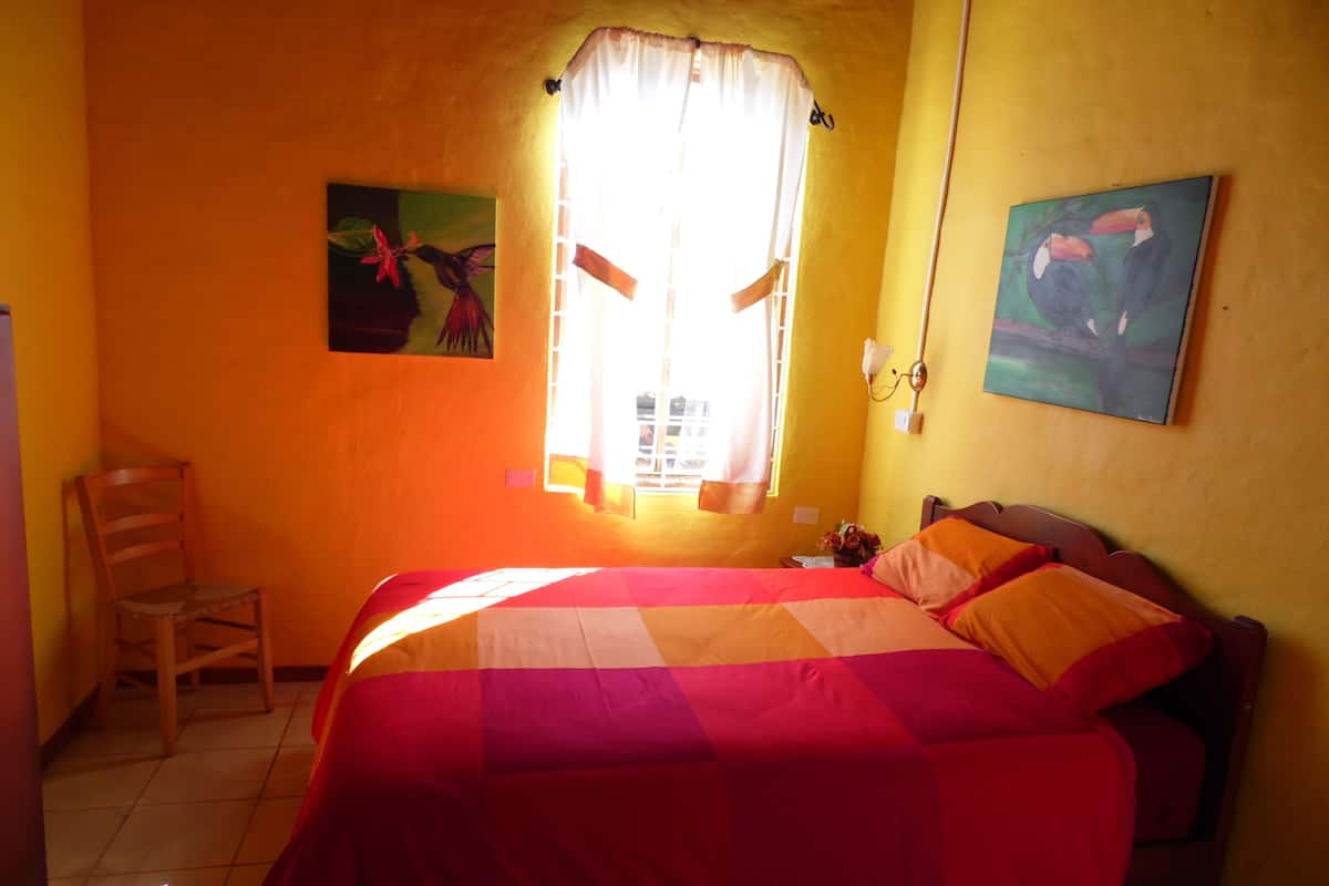 A room with a bed, a chair, a window with curtains, and two wall paintings.