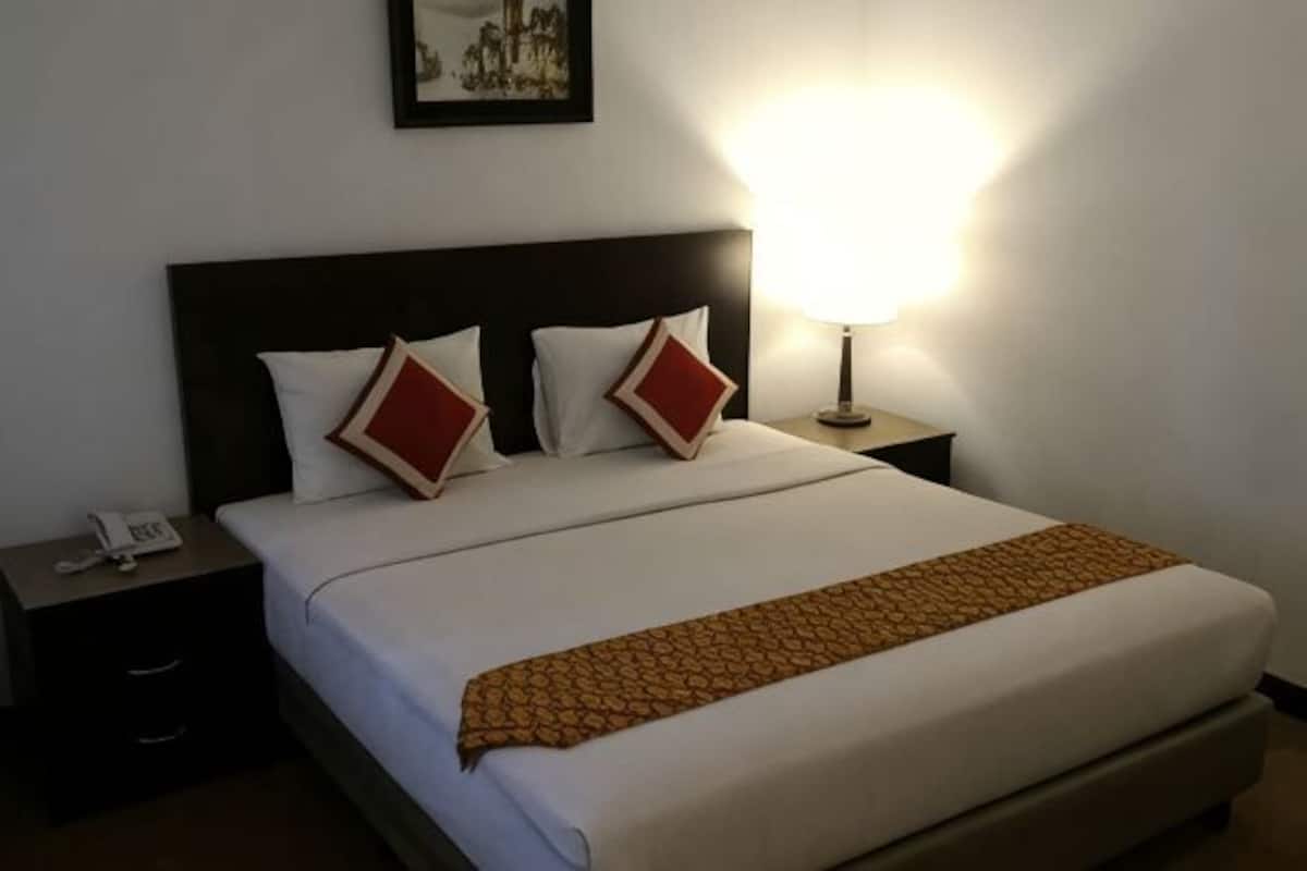 Una cama bien hecha con ropa blanca y dos almohadas rojas, una lámpara de noche y una mesita con un teléfono.