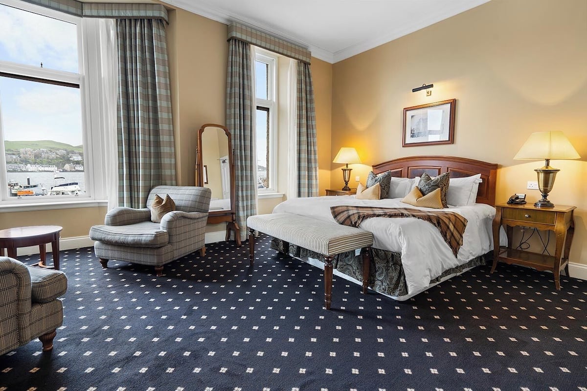 A hotel room with a bed, two armchairs, a bench, a nightstand, and a window with curtains.