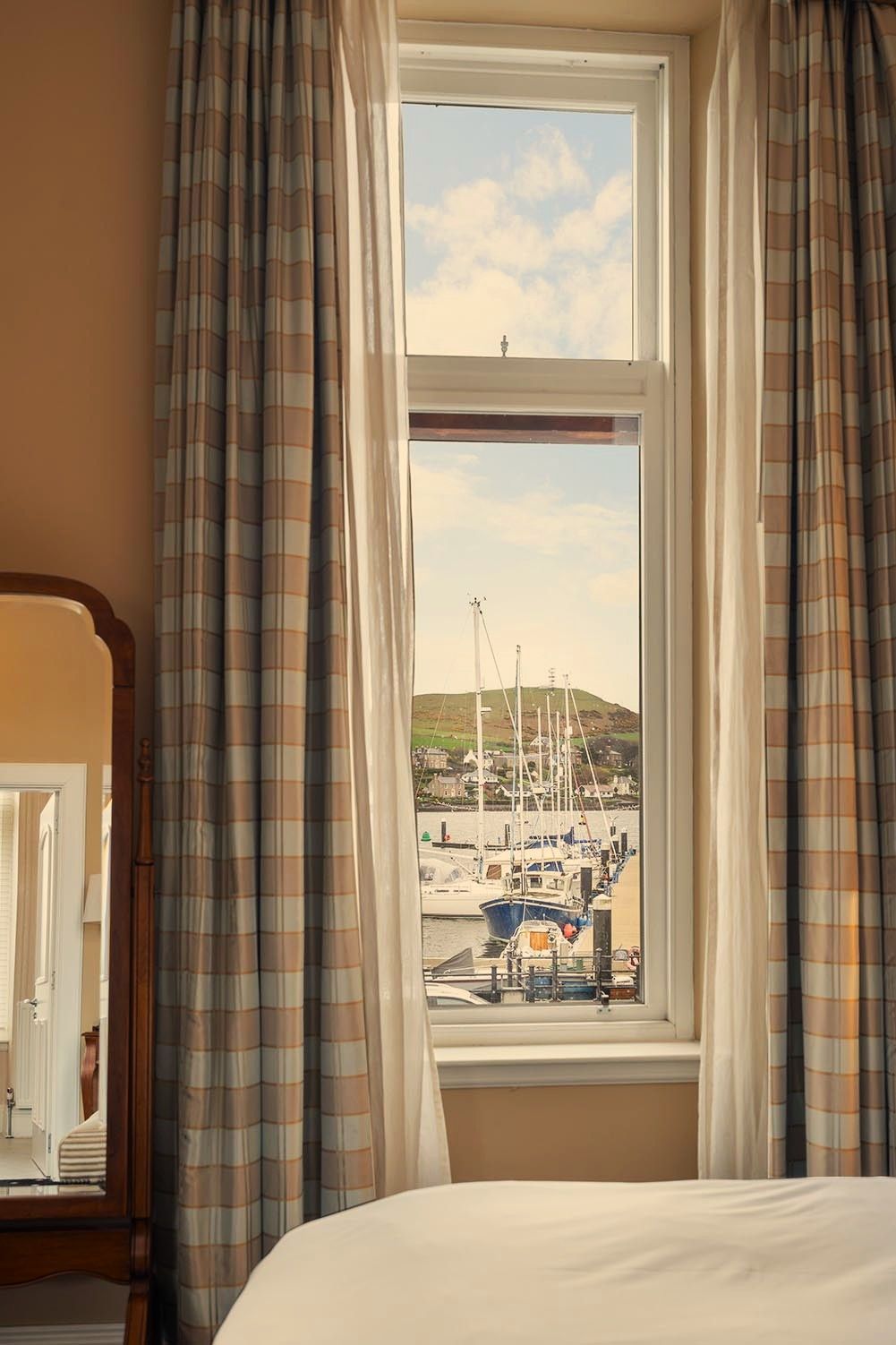 A bedroom with a large window, curtains, a bed, and a view of a marina with boats.