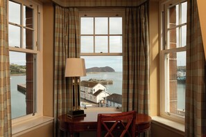 A room with a desk, chair, lamp, and a view of a coastal area with buildings and a pier. - The Royal Hotel Campbeltown  (Campbeltown)