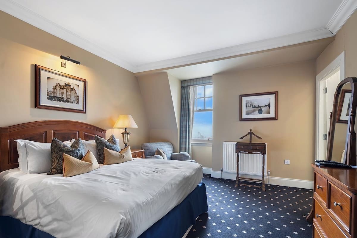 A hotel room with a large bed, a bedside table with a lamp, a chair, a dresser, and two framed pictures on the walls.