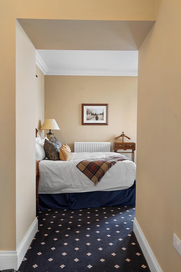 Habitación de hotel estrecha con cama, mesita de noche con lámpara, un cuadro en la pared y un radiador.