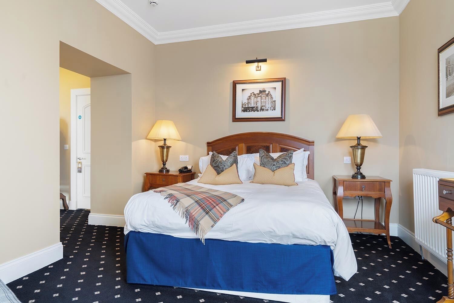A hotel room with a large bed, two bedside tables with lamps, a framed picture on the wall, and a radiator.