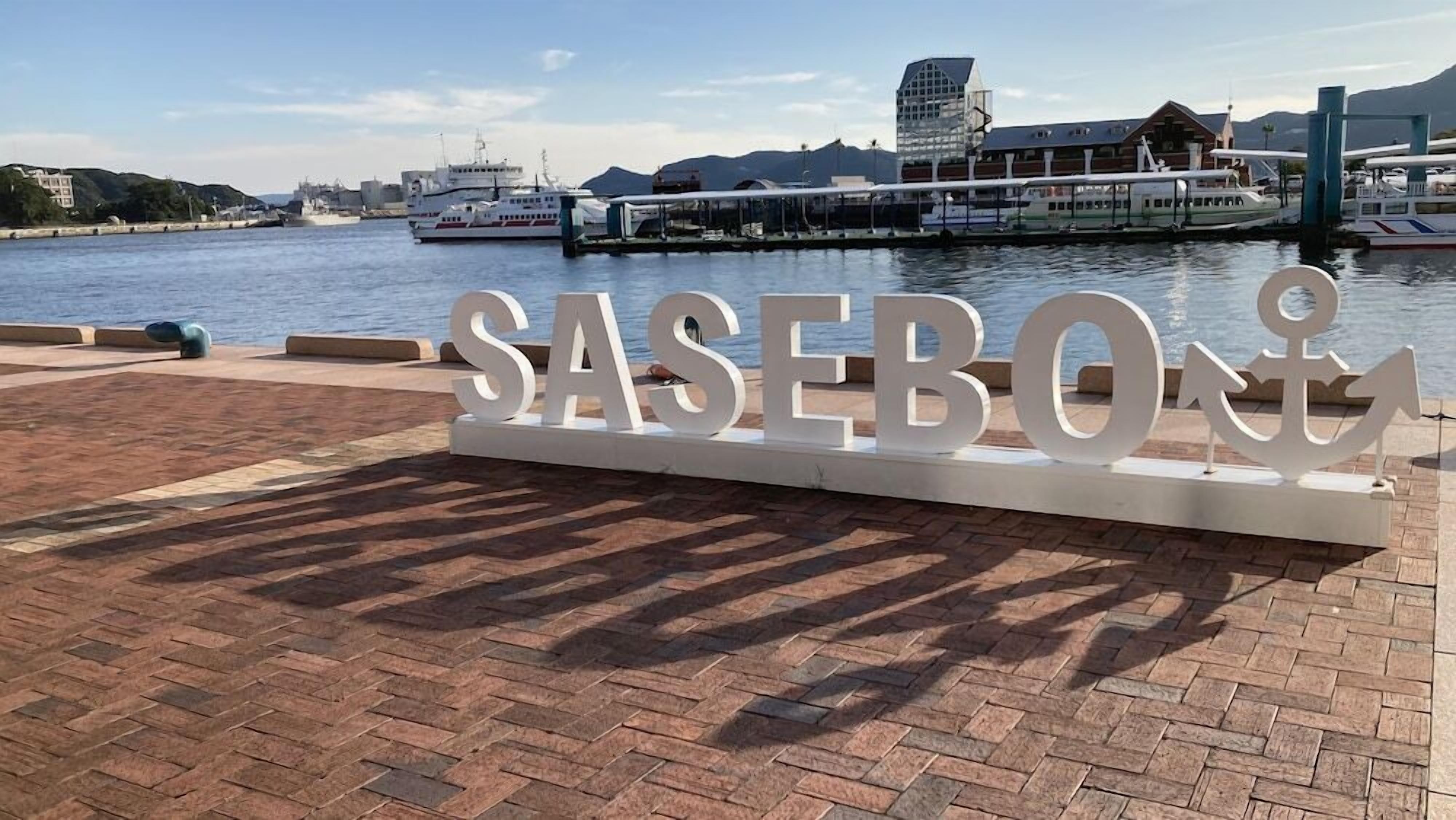 A large sign with the text "SASEBO" and an anchor symbol, located near a waterfront with boats and buildings in the background.