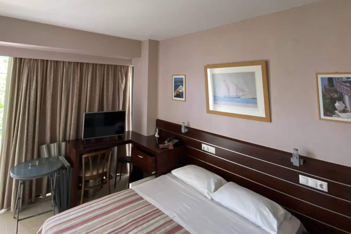 Une chambre d’hôtel avec un lit, un bureau équipé d’un ordinateur, une chaise et un téléviseur. Deux tableaux encadrés sont accrochés au mur.