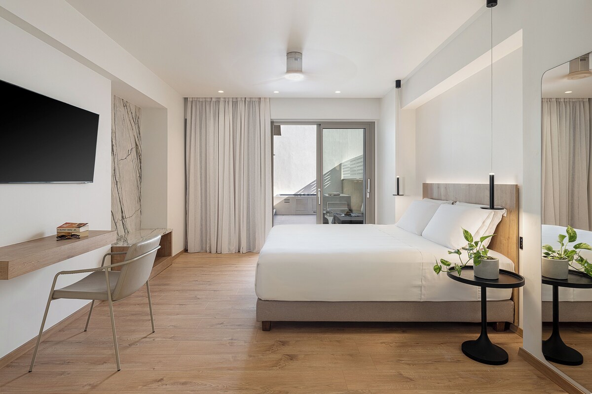 A modern hotel room with a bed, a desk, a chair, a small table with a lamp, and a TV mounted on the wall.
