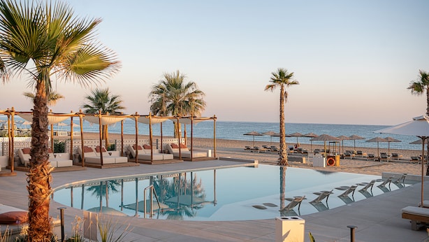 A poolside area with lounge chairs and umbrellas, a beach in the background, and a clear sky. - Iperion Beach (Rethymno)