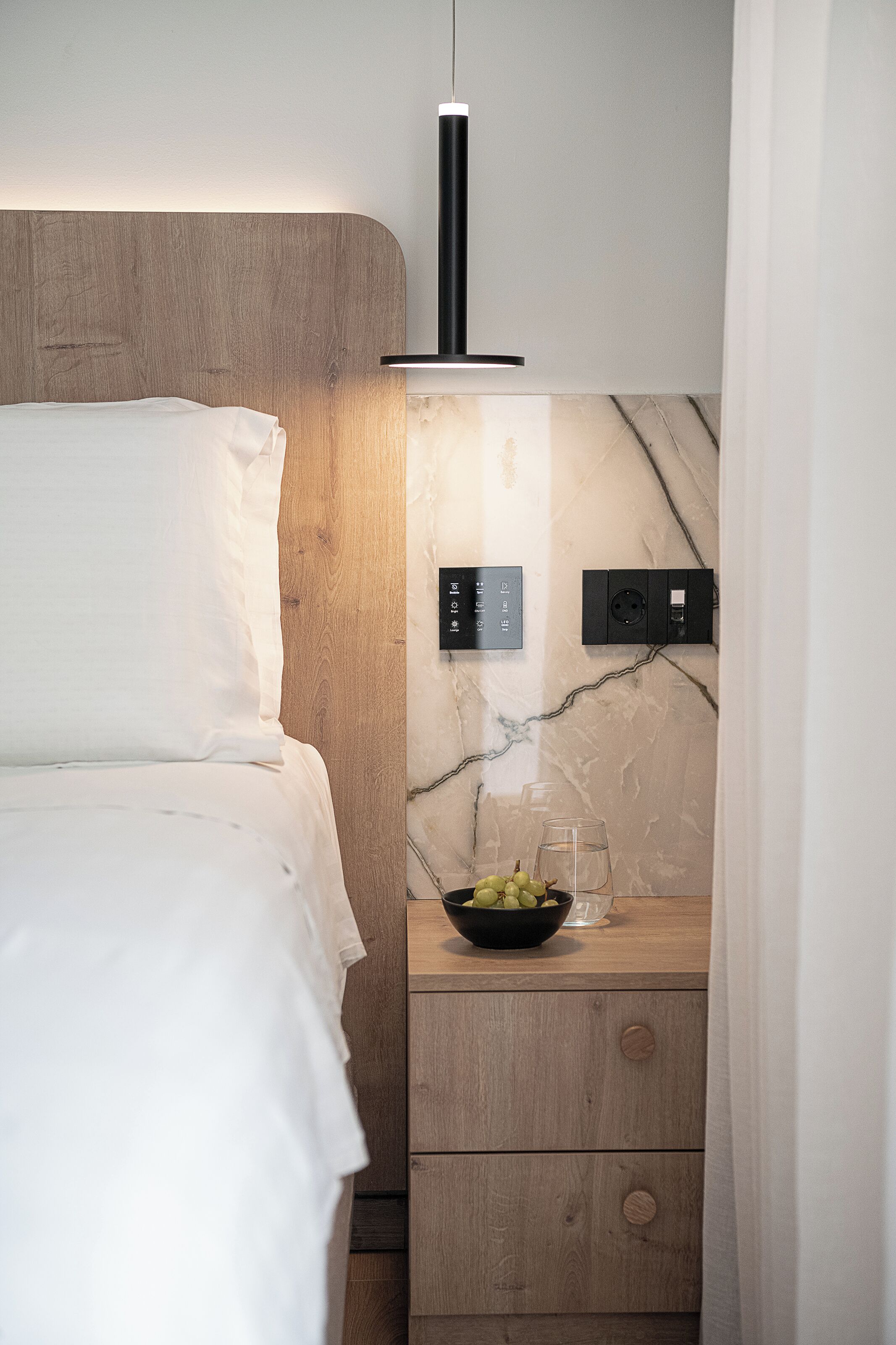 Quarto moderno com cabeceira de madeira, mesa de cabeceira com uma tigela de frutas e luminária fixada na parede.