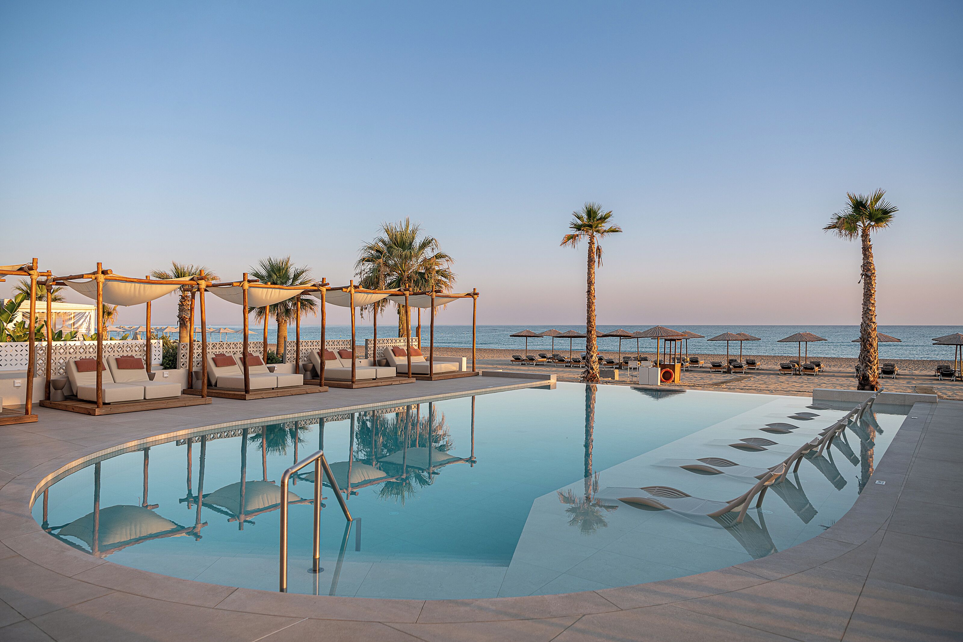 A poolside area with lounge chairs and umbrellas, a clear sky, and a calm sea in the background.