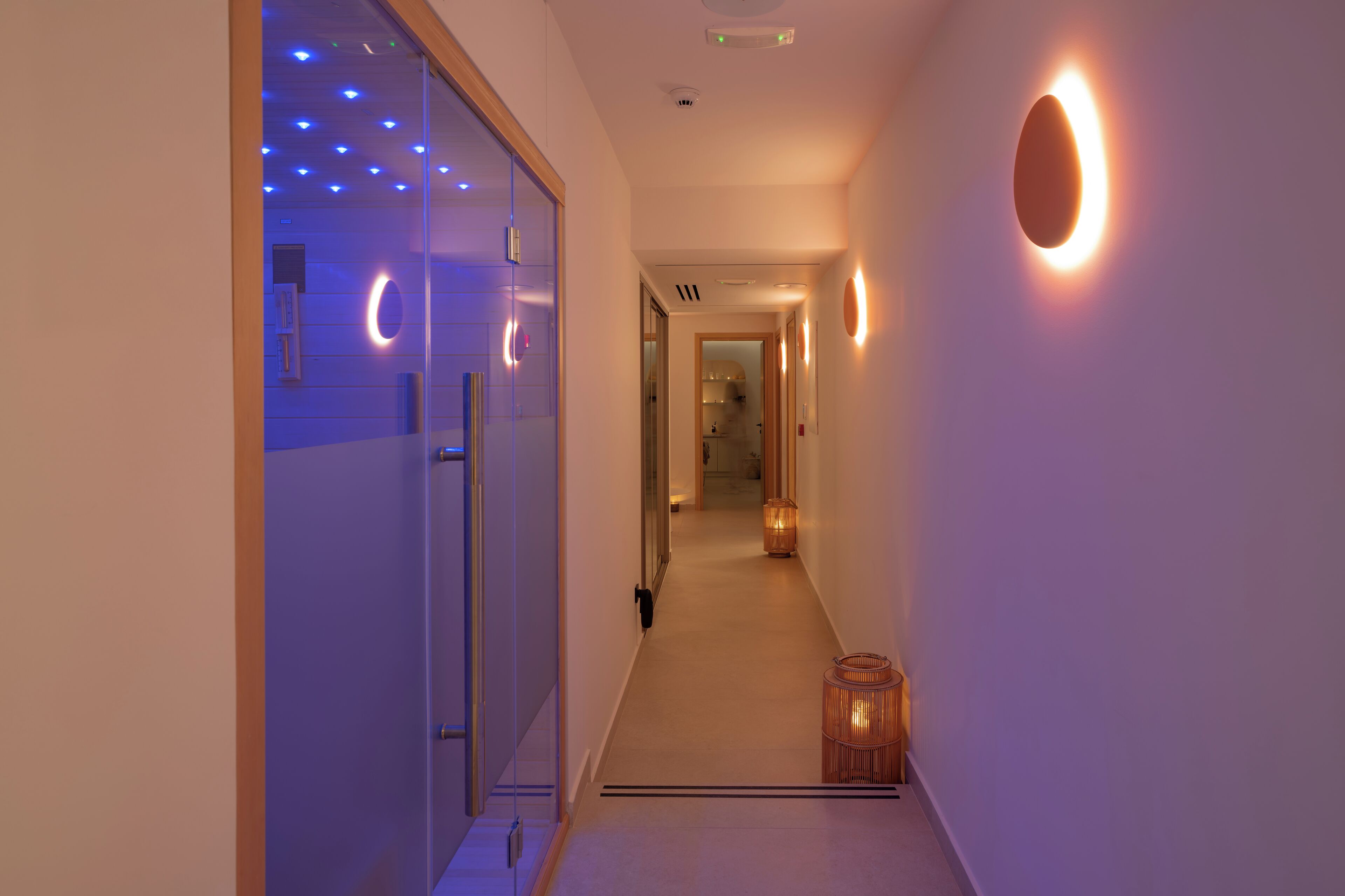 A hallway with a glass door on the left, a wooden door at the end, and a wall-mounted light fixture on the right.