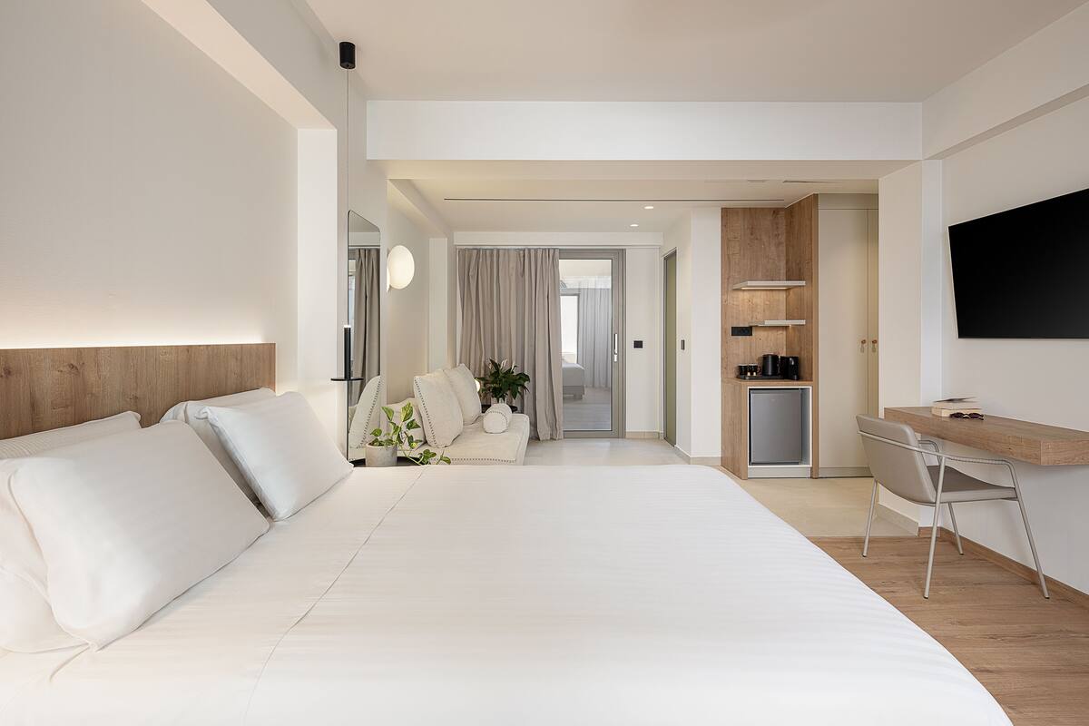 A modern hotel room with a large bed, a desk with a chair, a TV, and a kitchenette.
