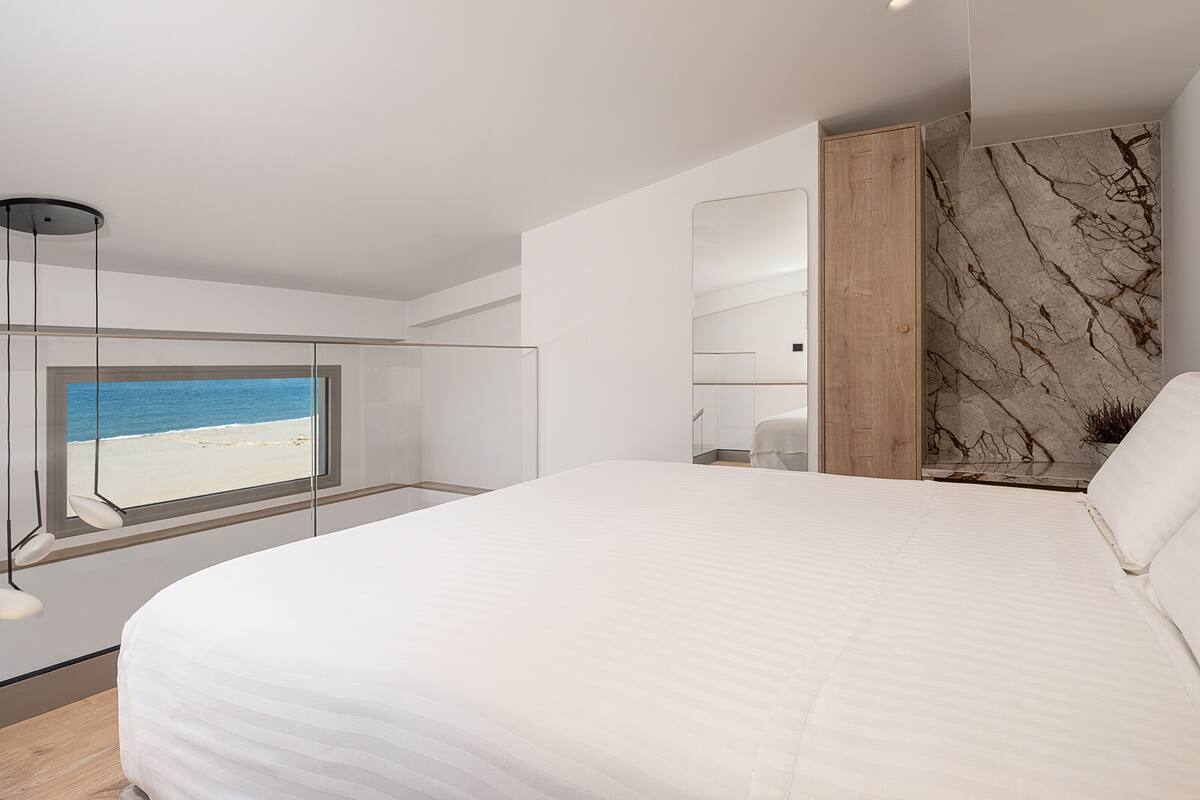 A modern bedroom with a large bed, a glass partition, a wooden wardrobe, and a view of the beach.