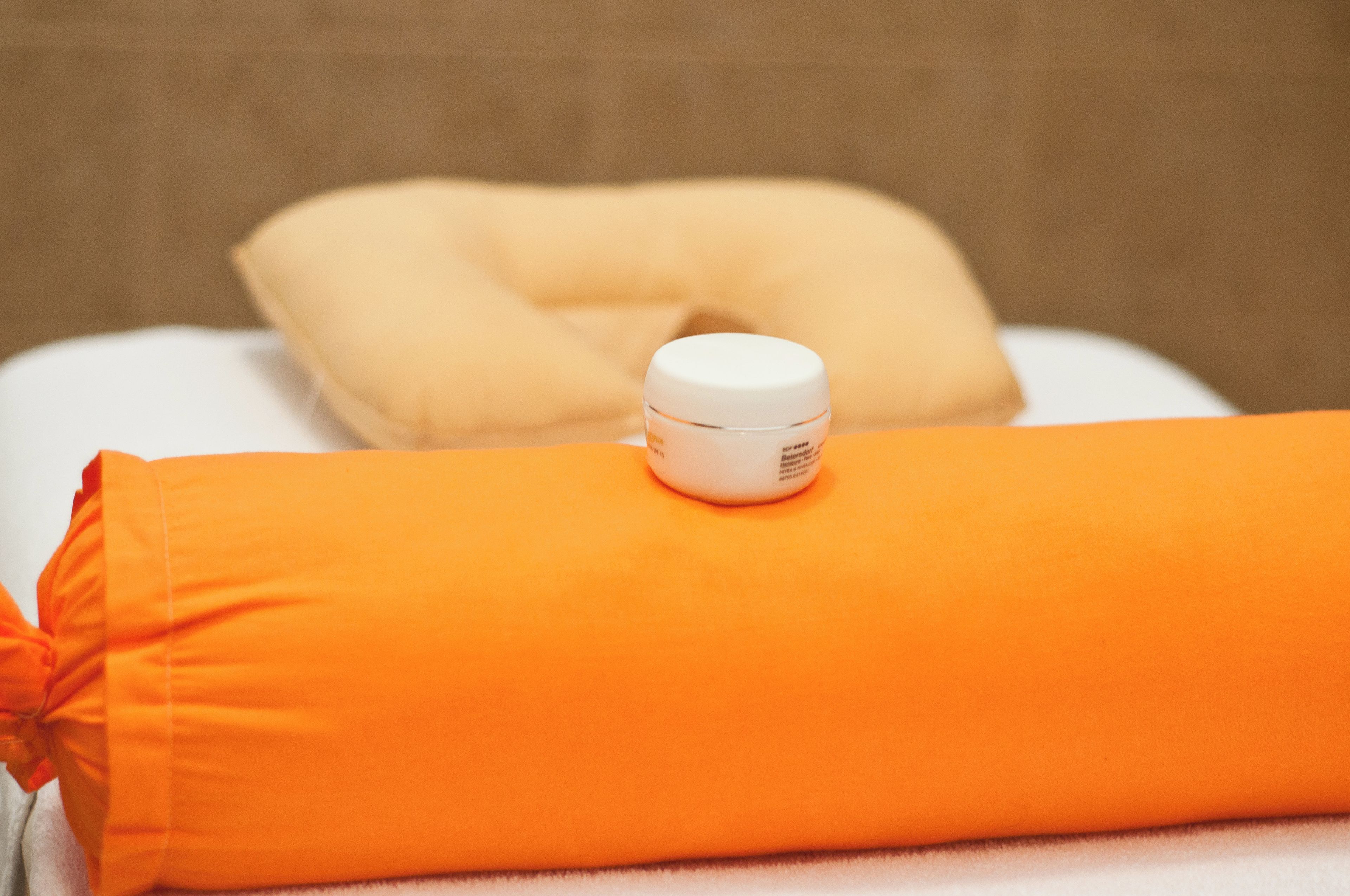 Un almohadón blanco y una toalla enrollada de color naranja sobre una cama.