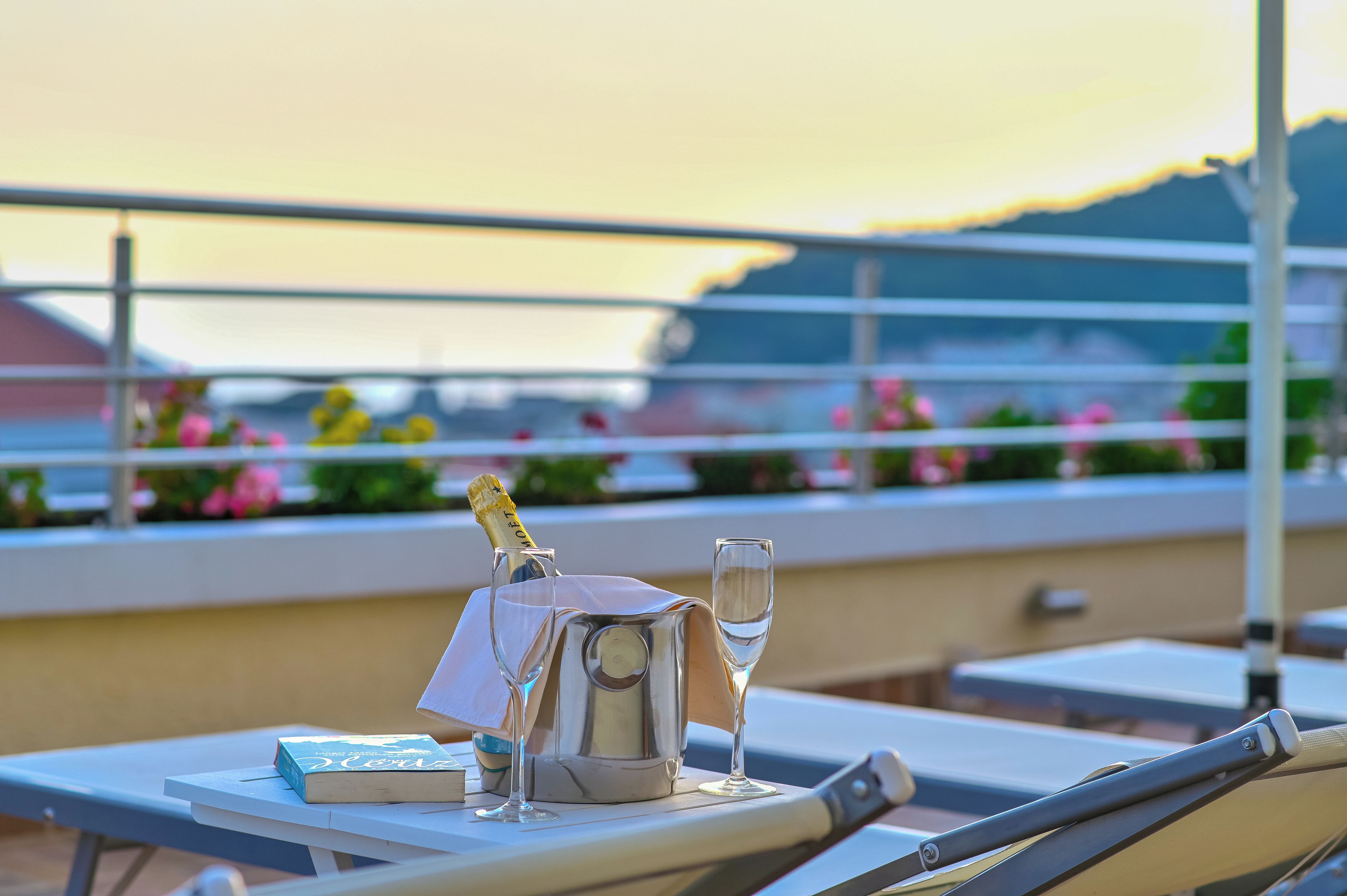 A table with a bottle of champagne, a glass, and a book on a terrace with a view of the sunset.