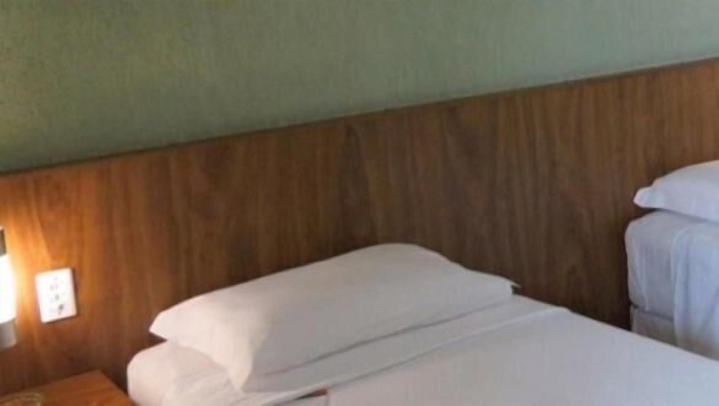Una habitación de hotel con cabecera de madera, una cama individual con ropa blanca y una mesita de noche con lámpara.