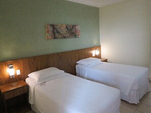 A hotel room with two single beds, wooden headboards, bedside lamps, and a small table with a glass container. - Steel Valley Hotel (Ipatinga)