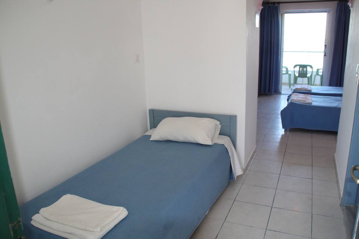 A simple room with two single beds, a tiled floor, and a view of a balcony with chairs.