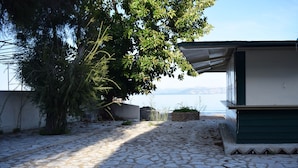 A stone-paved courtyard with a tree, a building with a green roof, and a view of the sea. - Akti Panela Beach Hotel (Corfu)