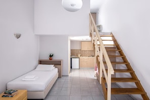 Ein Schlafzimmer mit einem Bett, Nachttisch und einer kleinen Pflanze. Eine Holztreppe führt zu einer Küchenzeile mit Schränken und einem Kühlschrank.