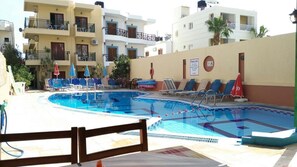 A swimming pool with lounge chairs and umbrellas, surrounded by multi-story buildings with balconies. - Stefania Studios (Hersonissos)
