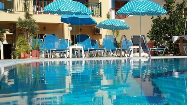 A swimming pool with blue chairs and umbrellas, surrounded by potted plants and a building with balconies.