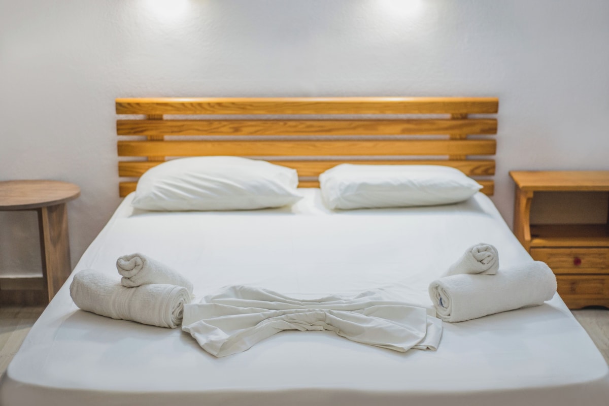 A neatly made bed with white linens and two pillows, a wooden headboard, and two bedside tables.