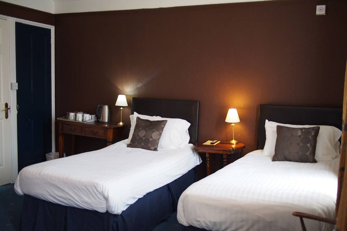 A hotel room with two single beds, a nightstand with a lamp, a desk with a coffee maker, and a door.