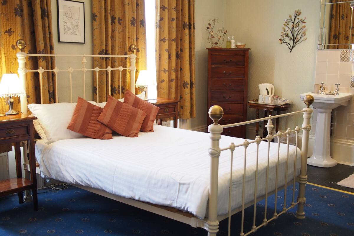 A neatly made bed with white linens and brown pillows, a wooden nightstand with a lamp, a dresser, and a bathroom with a sink and mirror.