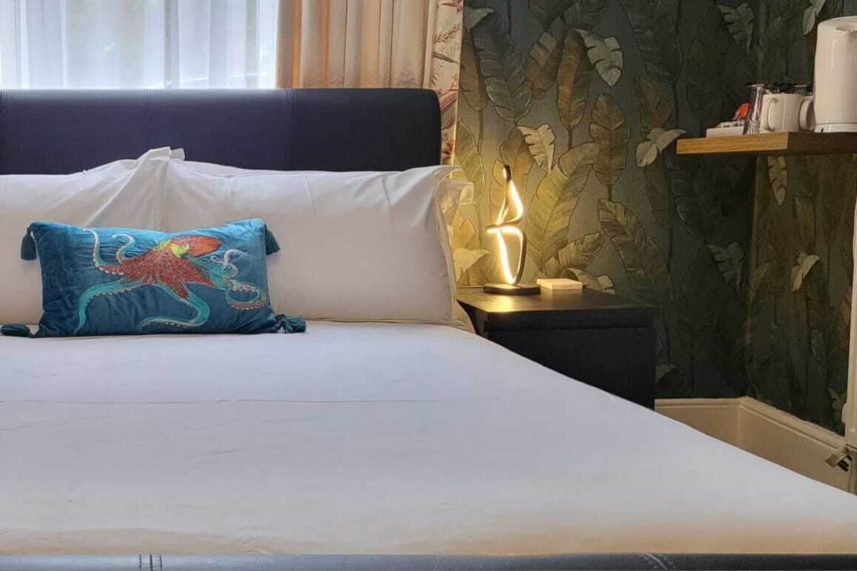 A bed with white linens, a blue pillow with a colorful design, two bedside tables with lamps, and a window with curtains.