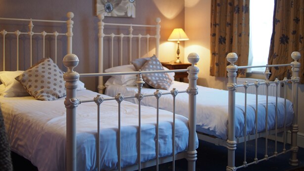 A double bed with a metal frame and white bedding, a bedside table with a lamp, and a floral painting on the wall. - The Coach House Canterbury (Canterbury)