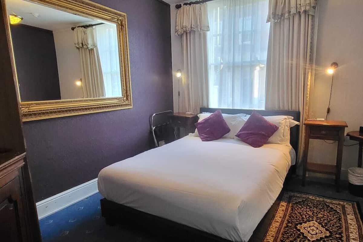 A bedroom with a bed, a dresser, a mirror, a bedside table, a radiator, and a window with curtains.