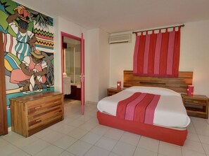 A hotel room with a large bed, a wooden dresser, a bathroom with a mirror, and a colorful mural on the wall. - Swalibo Hotel (Saint-Paul)