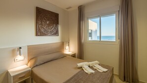 Une chambre d’hôtel avec un lit, une table de chevet, une œuvre d’art accrochée au mur, une fenêtre avec des rideaux et une vue sur la mer.