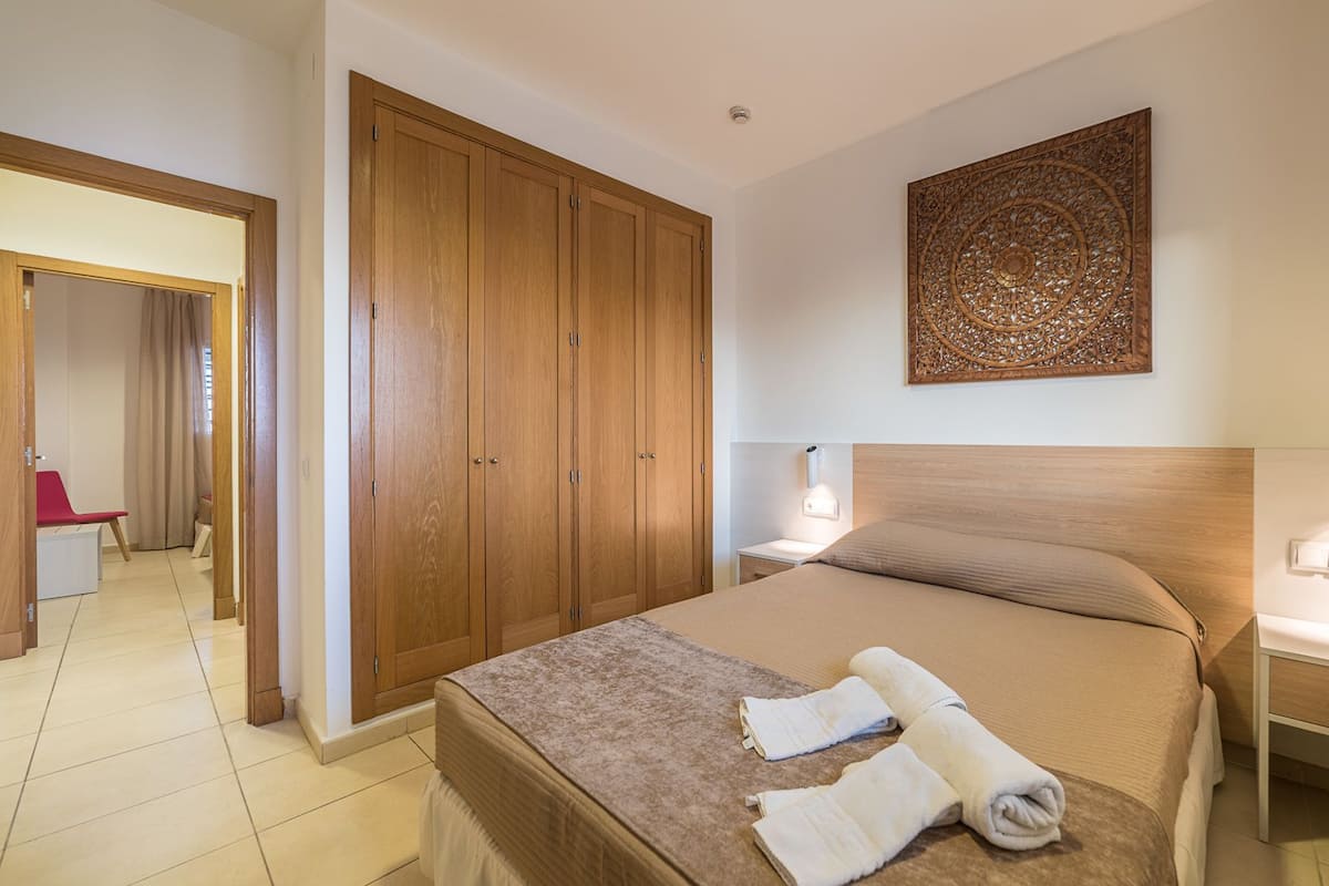 A hotel room with a large wooden wardrobe, a bed with beige bedding, a decorative wall piece, and a nightstand with a lamp.