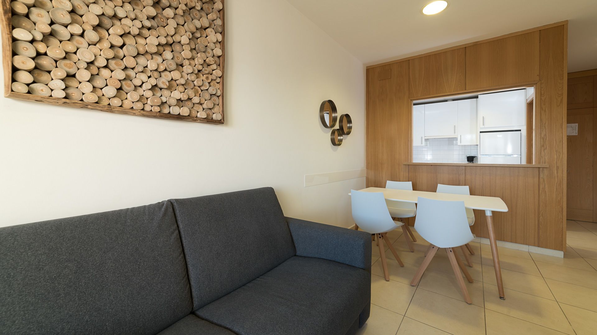 Una sala moderna con un sofá gris, una mesa de comedor con cuatro sillas y una zona de cocina con armarios de madera y un ventanal.