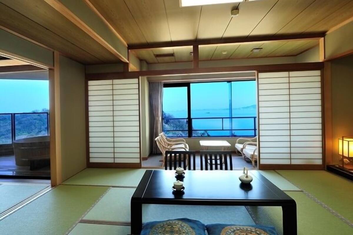 A traditional Japanese room with tatami flooring, a low table with tea set, and a view of the sea through large windows.