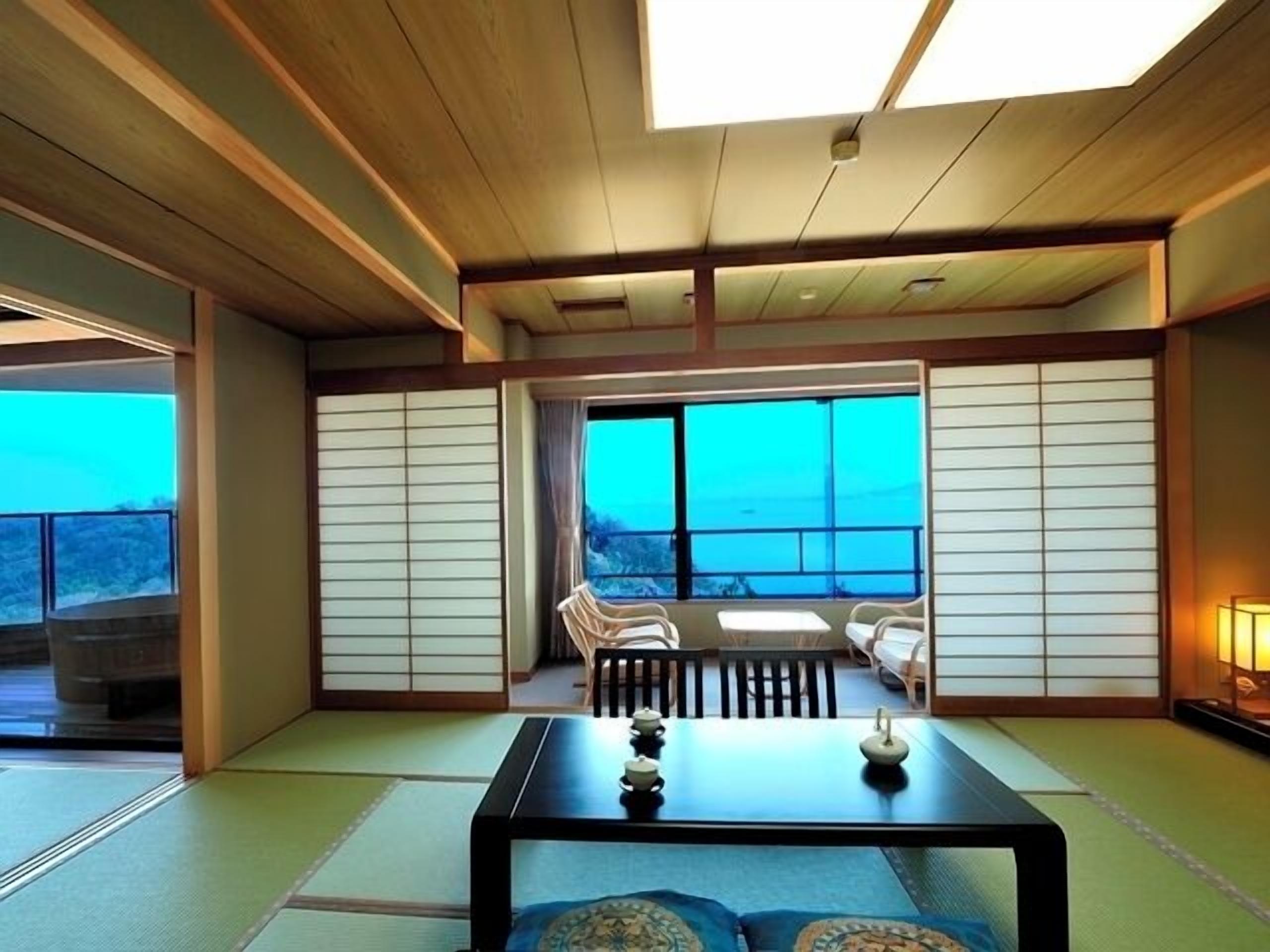 A traditional Japanese room with tatami flooring, a low table with tea sets, sliding doors, and a view through large windows.