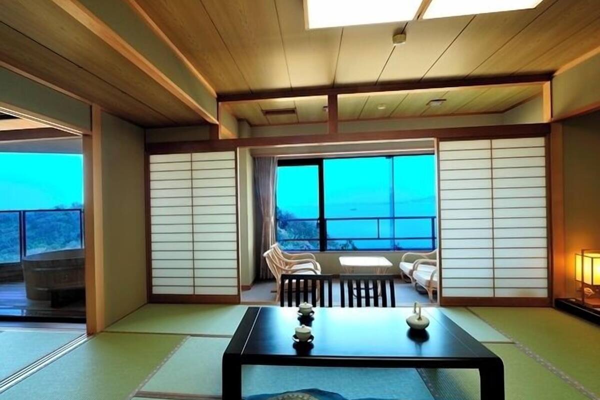 A traditional Japanese room with tatami flooring, a low table with tea sets, sliding doors, and a view through large windows.