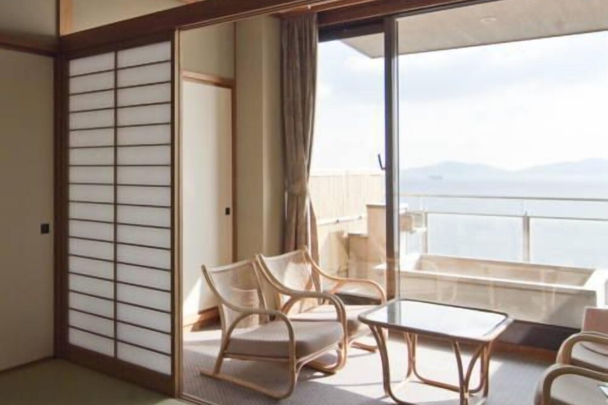 Una habitación con suelo de tatami, un panel corredizo shoji, un balcón con vistas y una mesa con sillas.